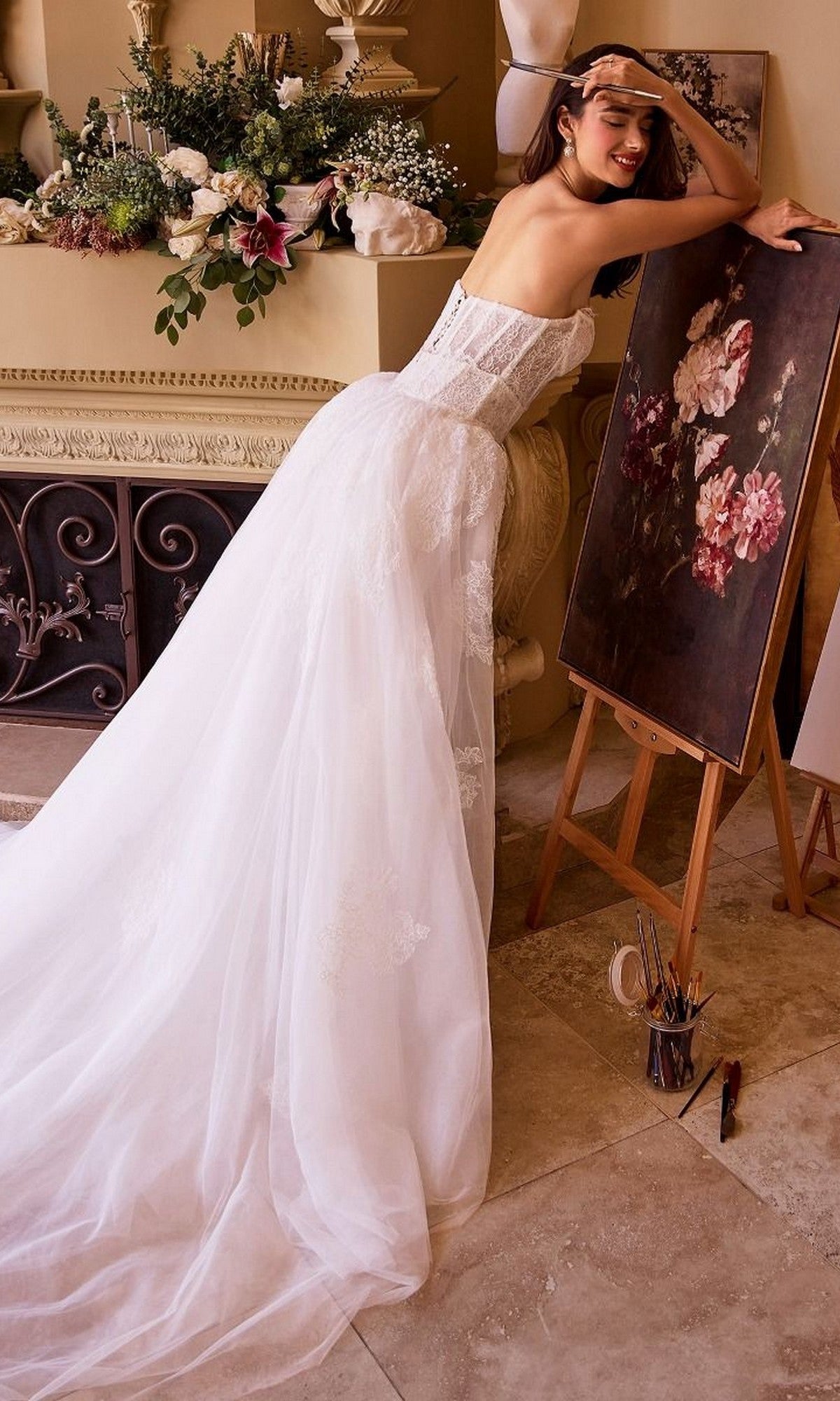 Dressed in the Andrea and Leo WL044 long formal dress, a woman leans beside an easel displaying a floral painting, surrounded by blossoms and art supplies in a warmly lit room—a sophisticated setting ideal for bridal celebrations.
